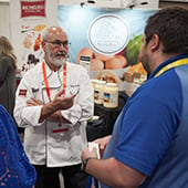 Man in chef's coat talking to another man in front of trade show booth